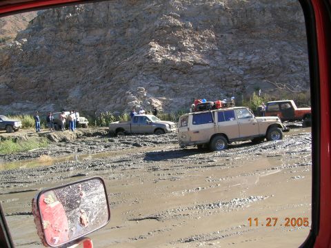 1985 Ford Bronco - Multiple vehicles navigate a muddy terrain with a rocky cliff face in the background.