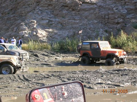 1985 Ford Bronco - Several off-road vehicles are stuck in a muddy terrain, with one orange Jeep Wrangler partially submerged.