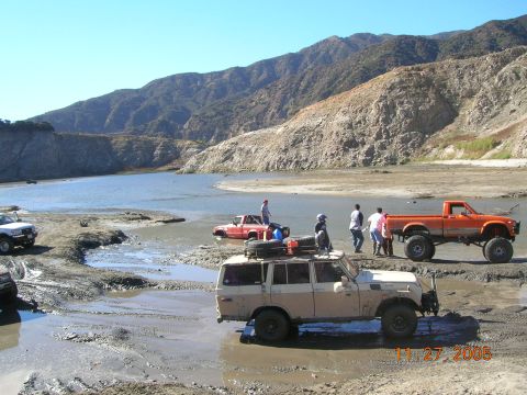 1985 Ford Bronco - Off-road vehicles navigate a muddy terrain by a body of water with a mountainous backdrop.