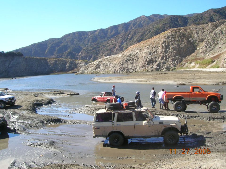 Several off-road vehicles are stuck in muddy water near a lake with mountains in the background. The date 11.27.2005 is visible