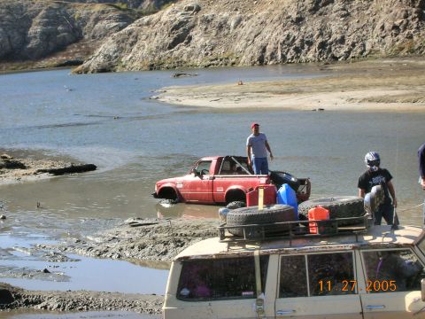 1985 Ford Bronco - Two off-road vehicles are partially submerged in muddy water near a rocky shoreline.