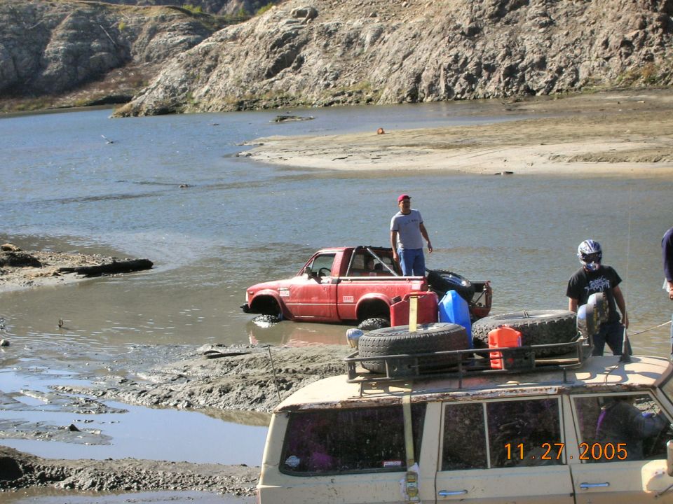 Red pickup truck stuck in muddy water, with a man standing in the bed. Another vehicle is in the foreground