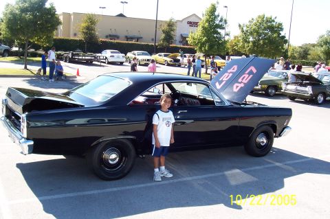 1969 Ford Ranchero - A black coupe with its hood open, displaying '427' in red lettering, stands in a parking lot with a child beside it.