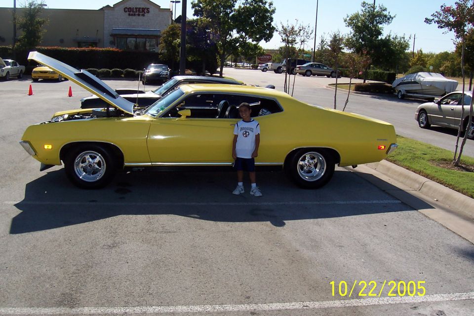 Yellow Ford fastback muscle car with hood up and polished five-spoke wheels at a car show