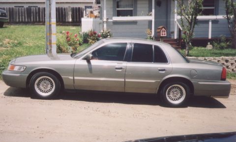 2000 Mercury Grand Marquis - Side profile of a silver 2000 Mercury Grand Marquis sedan parked on a dirt road in front of a house.
