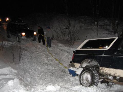 1992 Ford Bronco - Two SUVs are being pulled out of the snow at night with a yellow strap.