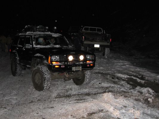 Black Jeep Cherokee XJ with snow on roof, modified bumper, and winch, in a snowy area at night