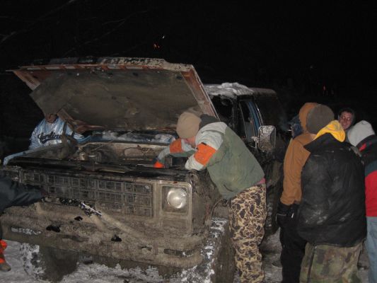 Mud-covered 1980s Ford Bronco with hood open, people working on engine in snowy conditions