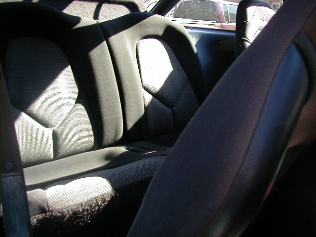 Dark fabric rear car seats with patterned inserts and a seatbelt. Another car is visible through the window