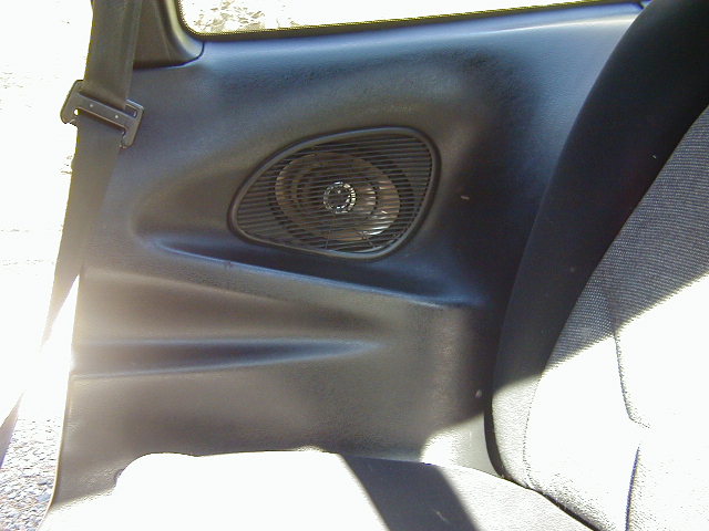 Car interior door panel with speaker grille and seat fabric visible. Seatbelt is also present
