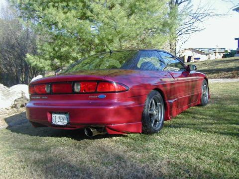 1994 Ford Probe - The rear of a red 1994 Ford Probe is visible, parked on a grassy area with trees and houses in the distance.