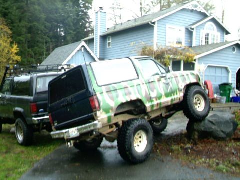 1987 Ford Bronco II - Two Ford Bronco II SUVs are parked outside a house, one is on rocks and has camouflage paint.