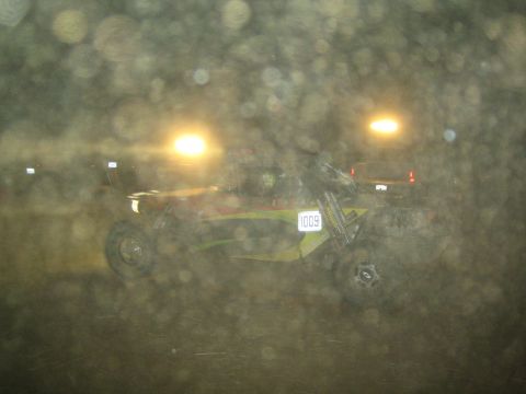 1990 Ford Bronco - green off-road race buggy numbered 1009 races over dirt at night, lit by floodlights and seen through a hazy, spotted foreground.