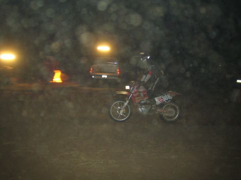 1990 Ford Bronco - helmeted rider on a dirt bike numbered 306 rides past a parked pickup truck at a night off-road event amid dust and lights.