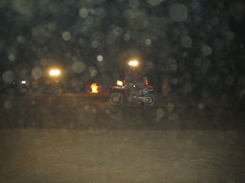 1990 Ford Bronco - motorcycle rider crosses a paved area at night with a small fire, floodlights and parked vehicles visible behind a low wall.
