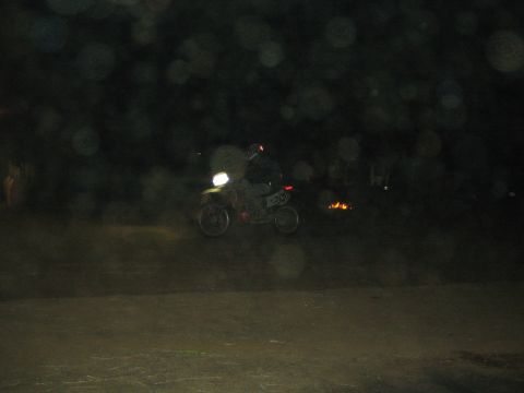 1990 Ford Bronco - dimly lit rider on a dirt bike with its headlight on crosses a dark dirt area as a small fire glows in the background.
