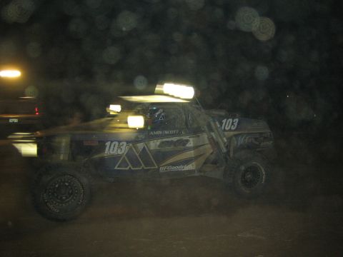 1990 Ford Bronco - blue and white off-road race truck numbered 103 with a mountain logo, BFGoodrich decals and a roof light bar races at night.