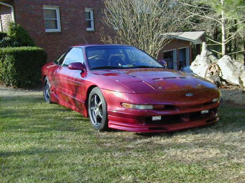 1994 Ford Probe - A red 1994 Ford Probe is parked on a grassy lawn in front of a house and trees.