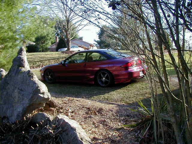 Red coupe parked on grass, three-quarter front view. Aftermarket wheels visible