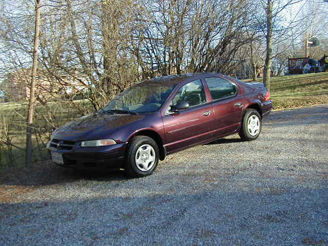 Maroon sedan parked on a gravel driveway next to bare trees. The car has a distinctive grille and headlight shape