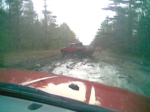 1976 GMC C/K Pickup - Two red pickup trucks on a muddy off-road trail surrounded by trees.