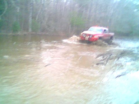 1976 GMC C/K Pickup - A red pickup truck navigates through a shallow, muddy body of water with trees in the background.