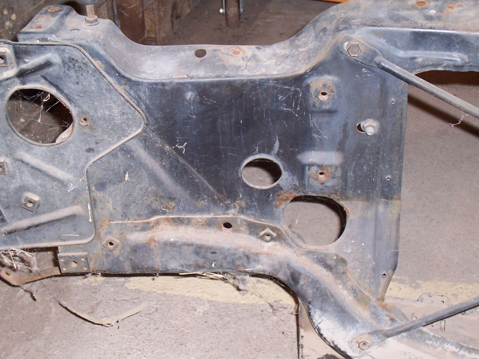 Bare rusty stamped-steel front subframe removed and lying on a concrete shop floor