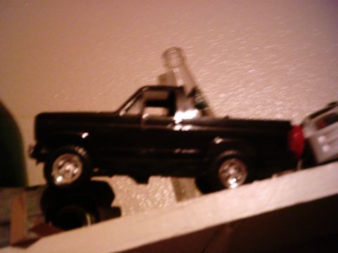 1986 Ford Bronco - A black toy truck is positioned on a shelf next to a white toy vehicle.