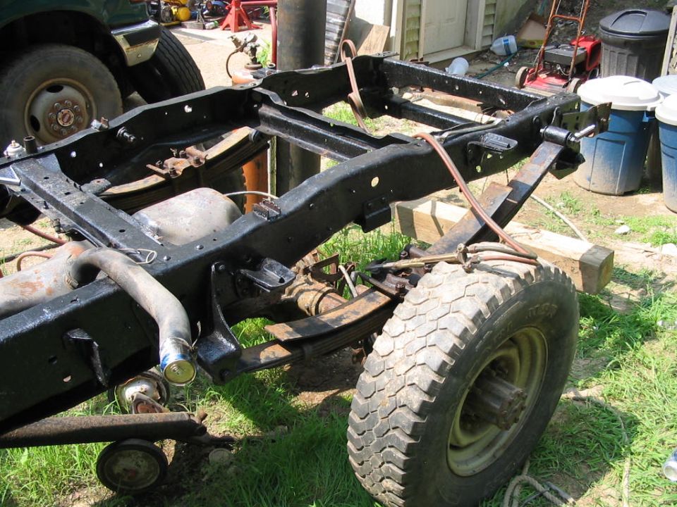 Stripped pickup frame on grass with leaf springs, fuel tank and one wheel