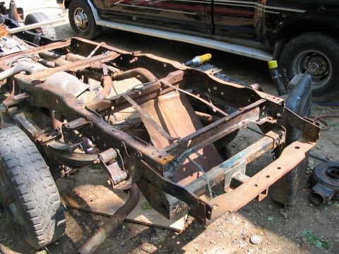 1985 Ford F-350 - The frame of a truck sits on the ground, showing significant rust and wear, with a black truck parked behind it.