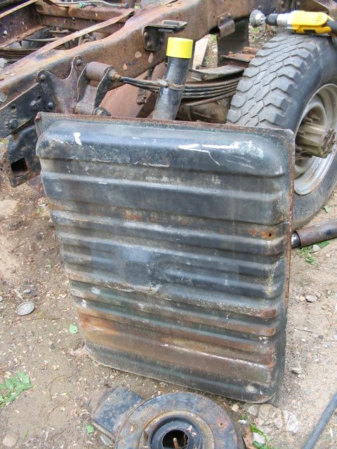 1985 Ford F-350 - A detached fuel tank with visible rust and wear, resting on the ground near a truck's chassis and tire.