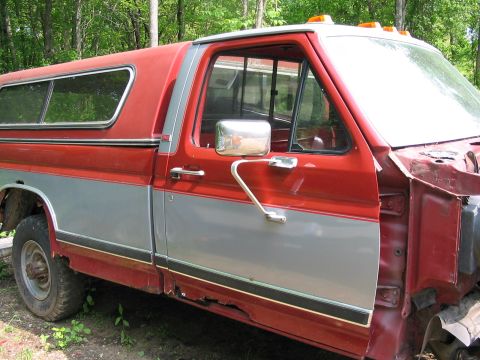 1985 Ford F-350 - A two-tone pickup truck with a camper shell is viewed from the side, revealing rust damage and missing body parts.
