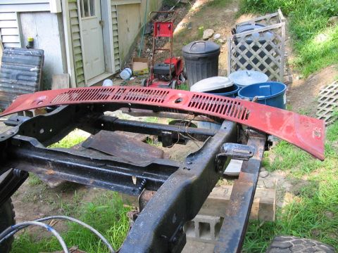 1985 Ford F-350 - The chassis of a truck is visible, with a red cowl panel resting on top. The surrounding area is cluttered with yard equipment and trash bi…