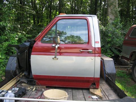 1985 Ford F-350 - A red and silver Ford F-Series truck cab sits on a wooden trailer, with another vehicle partially visible to the right.