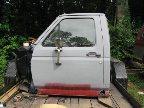 1985 Ford F-350 - A side view of a gray Ford F-Series truck cab with primer red lower body, showing rust damage.