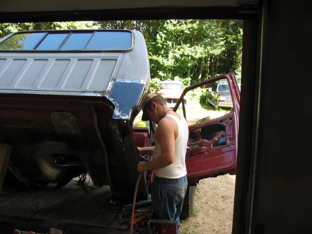 Man grinding rear cab corner of stripped red pickup cab with bare-metal roof in open shop doorway