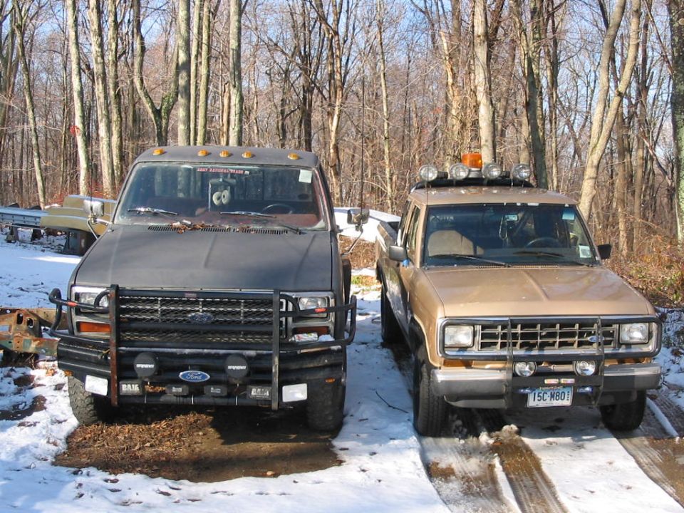 Two pickup trucks parked side-by-side in a snowy wooded area. The truck on the right has extra lights on its roof