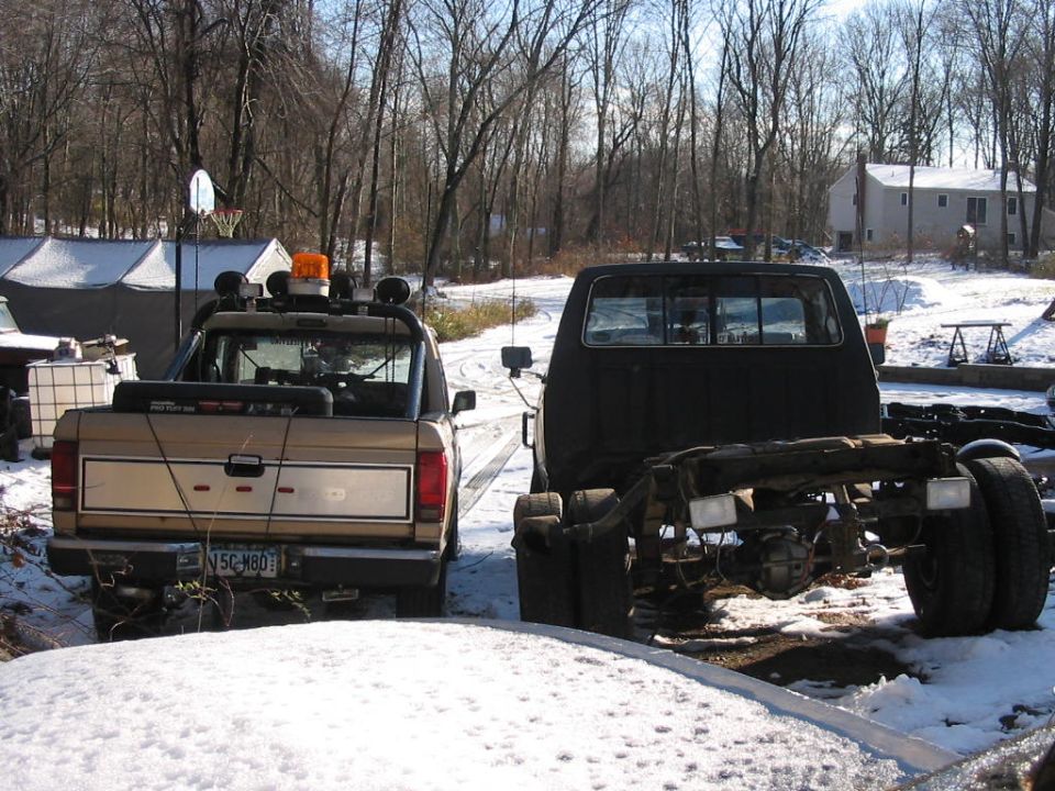 Rear view of a tan pickup truck next to a black truck chassis in a snowy yard