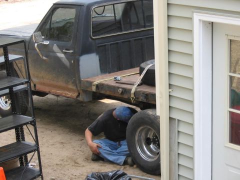 1985 Ford F-350 - A person works on the underside of a flatbed Ford pickup truck parked next to a building.