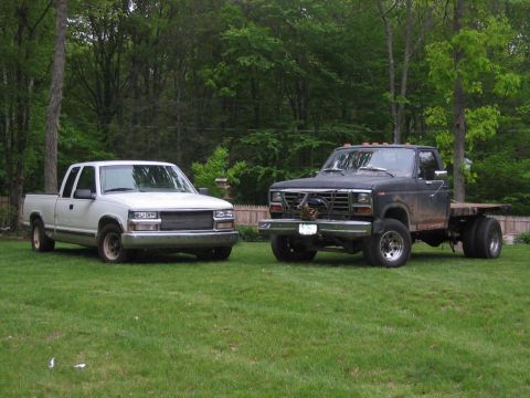 1985 Ford F-350 - Two pickup trucks, one black dually flatbed and one white standard pickup, are parked on a grassy lawn with trees in the background.