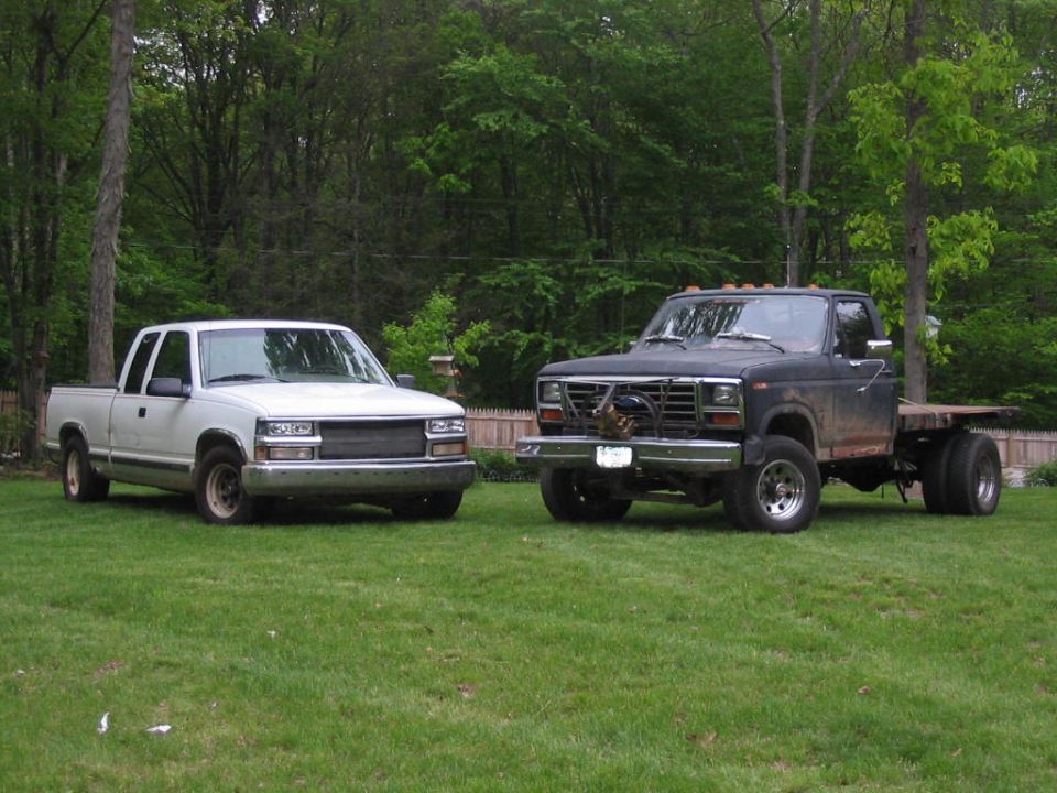 White GM extended-cab pickup beside a black Ford flatbed dually with brush guard and winch on lawn