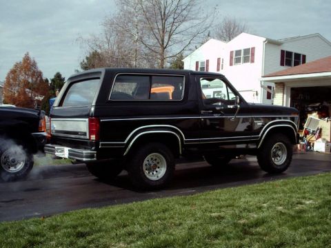1984 Ford Bronco - A black Ford Bronco is parked on a driveway, with another vehicle emitting smoke beside it.