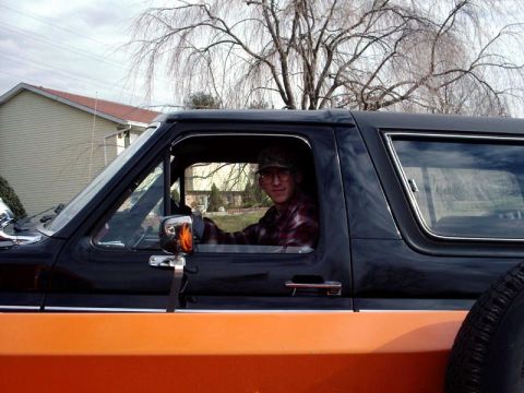 1984 Ford Bronco - A man sits behind the wheel of a two-tone black and orange Ford Bronco, likely from the 1980s.