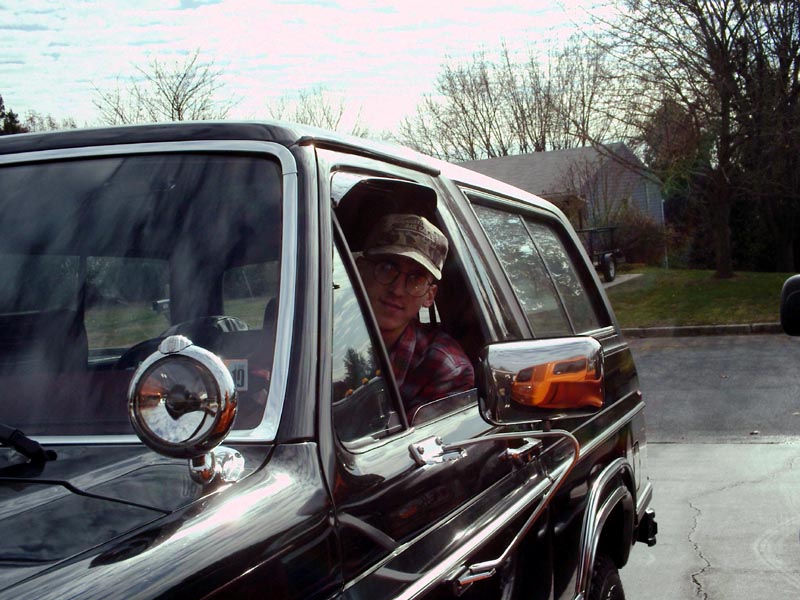 Person wearing a hat and glasses looks out the driver's window of a dark-colored Ford Bronco
