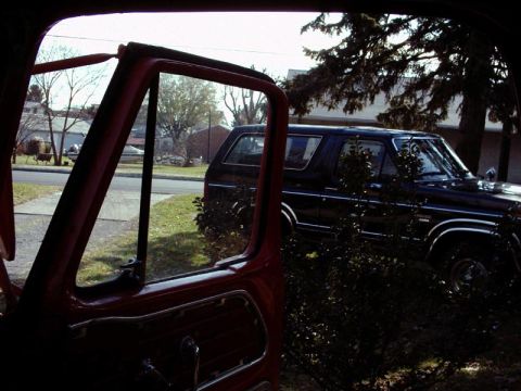 1984 Ford Bronco - A dark blue SUV is parked on a residential street, partially obscured by bushes and trees.