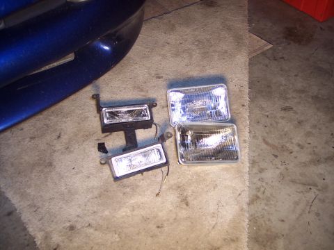 1995 Ford Probe - Four headlights are laid out on a concrete floor, with a portion of a blue car's bumper visible in the upper left corner.