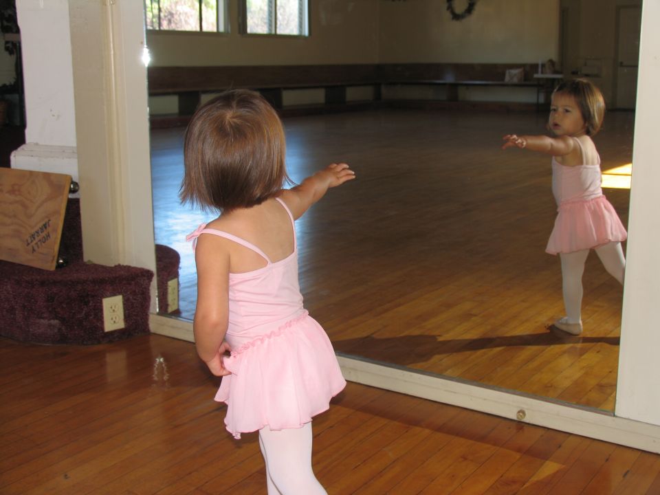 Young child in pink tutu and tights reaches towards a mirror in a dance studio