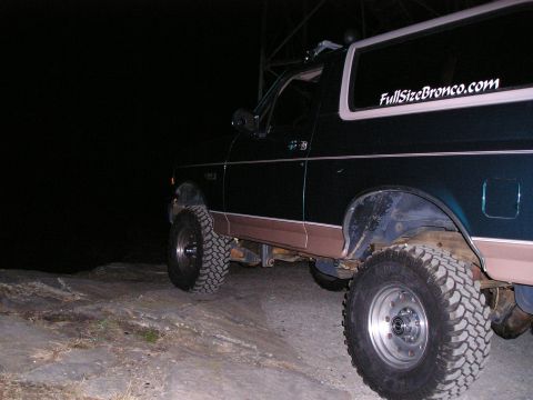 1996 Ford Bronco - A dark-colored, lifted Ford Bronco is parked on a rocky surface at night, with a website sticker on the rear window.