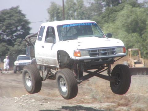 1984 Ford Bronco - A lifted white off-road truck with large tires is airborne, with another vehicle visible in the background.