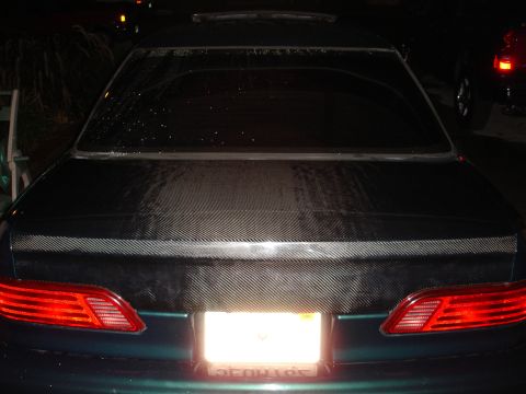 1993 Ford Taurus - The rear of a dark green car features a carbon fiber trunk lid and horizontal taillights, with a license plate obscured by glare.
